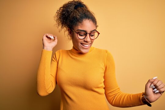 Young beautiful african american girl wearing sweater and glasses over yellow background Dancing happy and cheerful, smiling moving casual and confident listening to music