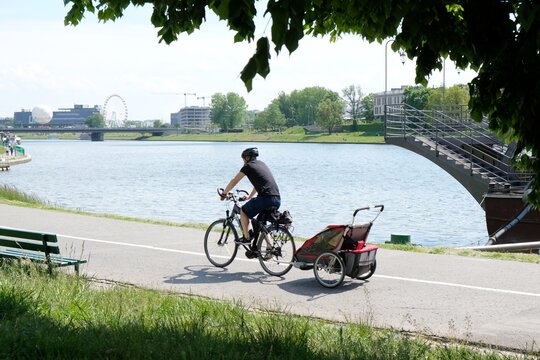 A Man Is Ridding A Bicycle With A Trailer For Child Along The Bike Path Along The Vistula River. Cracow, Poland