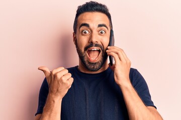 Young hispanic man having conversation talking on the smartphone pointing thumb up to the side smiling happy with open mouth