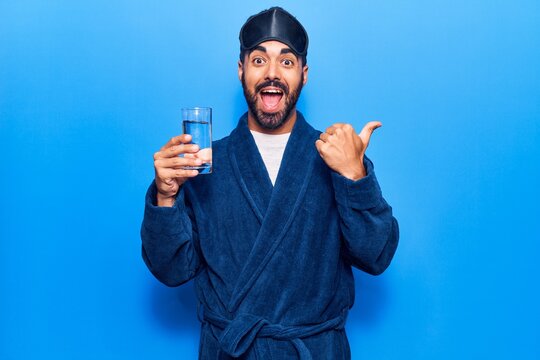 Young hispanic man wearing sleep mask and robe drinking water pointing thumb up to the side smiling happy with open mouth