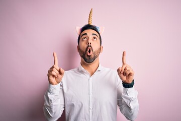 Young handsome man with beard wearing funny unicorn diadem over pink background amazed and surprised looking up and pointing with fingers and raised arms.