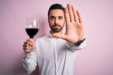 Young handsome man with beard drinking glass of red wine over isolated pink background with open hand doing stop sign with serious and confident expression, defense gesture