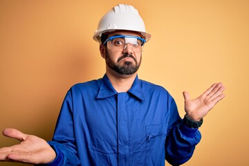 Mechanic man with beard wearing blue uniform and safety glasses over yellow background clueless and...