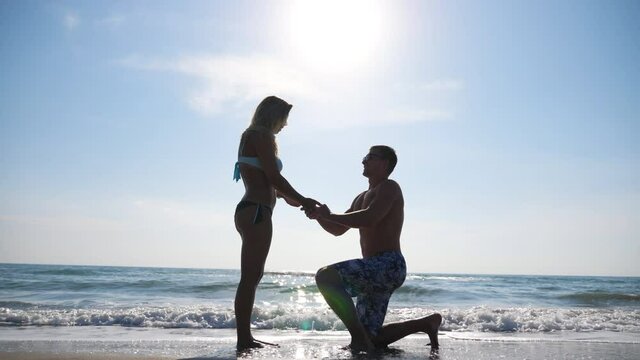 Young man stand on knee and marriage proposal to his girlfriend summer vacation. Happy pair hugging and kissing on seashore. View on newlyweds at seascape background. Concept of love and happiness