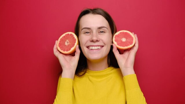 Funny crazy playful young woman in yellow sweater making silly monkey face, squinting eyes, bulging ears with grapefruit, comic girl having fun of citrus fruit, isolated on red studio background