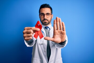 Young handsome doctor man with beard wearing stethoscope holding red hiv ribbon with open hand...