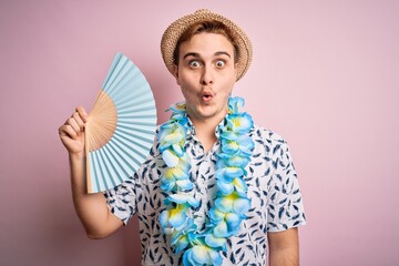 Young handsome redhead tourist man on vacation wearing hawaiian lei and hat using hand fan scared...