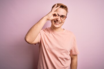 Young handsome redhead man wearing casual t-shirt standing over isolated pink background doing ok gesture with hand smiling, eye looking through fingers with happy face.