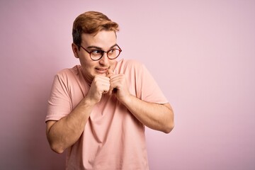 Young handsome redhead man wearing casual t-shirt standing over isolated pink background laughing nervous and excited with hands on chin looking to the side