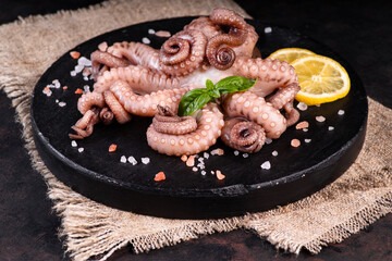 Raw octopus tentacles on a black round plate on a black background with slices of lemon and green...