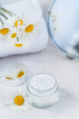 Fototapeta na wymiar Herbal cosmetic cream in opened glass container and fresh chamomile flowers, towel, mirror on a white background. Natural organic moisturizer skincare product. Selective focus. Vertical. Copy space.