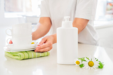 Obraz na płótnie Canvas Eco friendly cleaning dish soap with natural chamomile flower ingredient and woman putting clean white cups and plates on white kitchen table. Skincare for housekeeping. Non-toxic cleaning supplies.