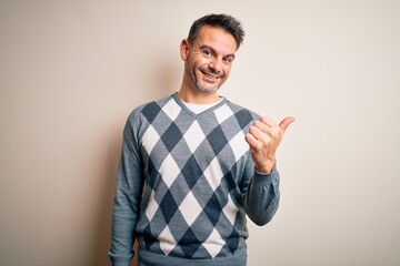 Fototapeta premium Young handsome man wearing casual sweater standing over isolated white background smiling with happy face looking and pointing to the side with thumb up.