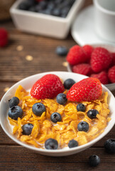 Corn flakes with blueberries and raspberries in white bowl in dark wooden desk.