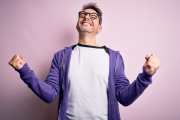 Young handsome man wearing purple sweatshirt and glasses standing over pink background very happy...