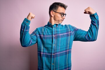 Young handsome man wearing casual shirt and glasses standing over isolated pink background showing arms muscles smiling proud. Fitness concept.