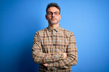 Obraz premium Young handsome man wearing casual shirt and glasses standing over blue background with serious expression on face. Simple and natural looking at the camera.