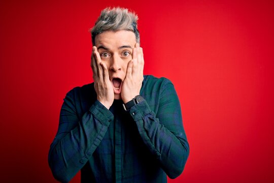 Young handsome modern man wearing elegant green shirt over red isolated background afraid and shocked, surprise and amazed expression with hands on face