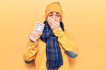 Young handsome bald man wearing winter clothes holding pills covering mouth with hand, shocked and...