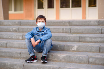 Schoolboy in protective mask sits on stairs outdoors in schoolyard. Pandemic coronavirus 2020.