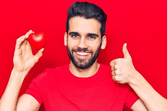 Young handsome man with beard holding red heart smiling happy and positive, thumb up doing excellent and approval sign