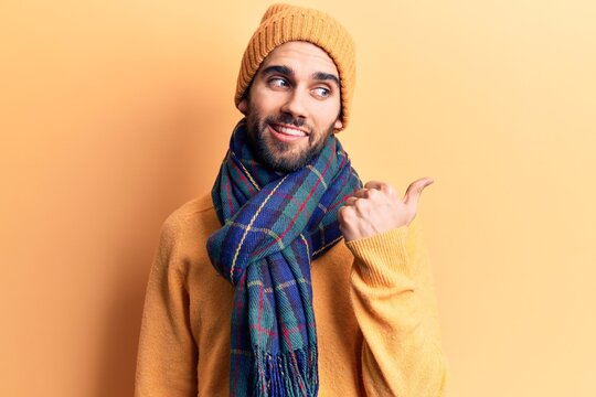Young handsome man with beard wearing wool cap and scarf smiling with happy face looking and pointing to the side with thumb up.