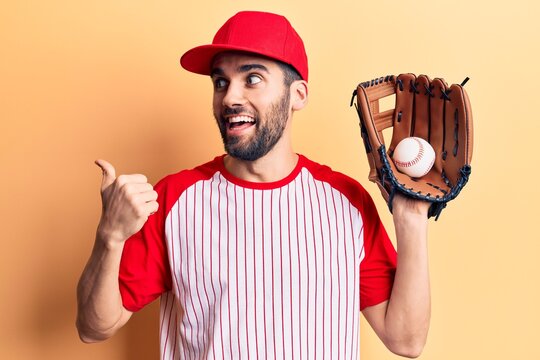 Young handsome man with beard playing baseball using ball and glove pointing thumb up to the side smiling happy with open mouth