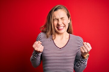 Young beautiful blonde woman wearing casual striped t-shirt over isolated red background excited for success with arms raised and eyes closed celebrating victory smiling. Winner concept.