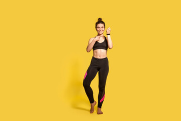 Fototapeta na wymiar Beautiful happy athletic woman with hair bun in tight sportswear pointing at wristwatch, showing fitness tracker, electronic gadget application for sport trainings. full length studio shot, isolated