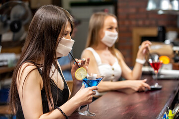 girls in medical masks chatting in a pub. girls in medical masks are talking at the bar. girls in medical masks drink cocktails