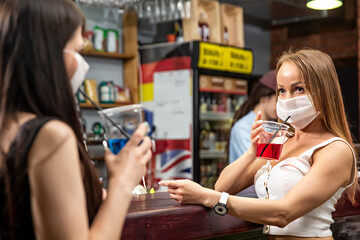 girls in medical masks chatting in a pub. girls in medical masks are talking at the bar. girls in medical masks drink cocktails