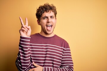 Fototapeta premium Young blond handsome man with curly hair wearing casual striped sweater smiling with happy face winking at the camera doing victory sign. Number two.