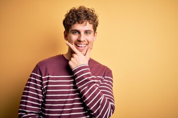 Young blond handsome man with curly hair wearing casual striped sweater looking confident at the camera smiling with crossed arms and hand raised on chin. Thinking positive.