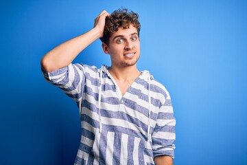 Young blond handsome man with curly hair wearing casual striped sweatshirt confuse and wonder about question. Uncertain with doubt, thinking with hand on head. Pensive concept.