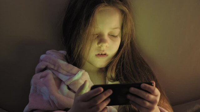 Child insomnia. Late night movie. Tired little girl watching disturbing video on phone in bed.