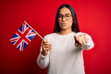 Young asian woman holding united kingdom flag for brexit referendum over red background pointing with finger to the camera and to you, hand sign, positive and confident gesture from the front