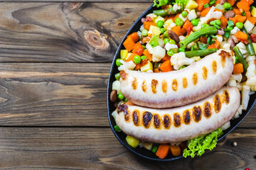 Grilled sausage with sauteed vegetables in cast iron skillet. Top view, wooden background.
