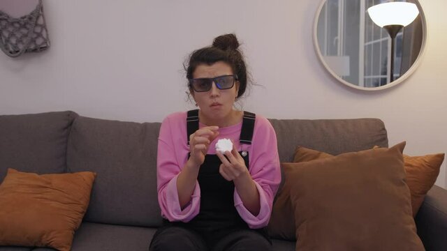 front view of woman in 3d glasses sitting on sofa and eating zefir while watching tv in living room