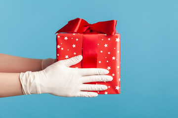 Profile side view closeup of human hand in white surgical gloves holding red gift box. sharing, giving or delivery concept. indoor, studio shot, isolated on blue background.