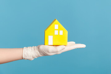 Fototapeta na wymiar Profile side view closeup of human hand in white surgical gloves holding yellow paper house exterior in hand. indoor, studio shot, isolated on blue background.