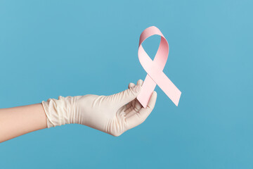 Profile side view closeup of human hand in white surgical gloves shwoing and holding pink ribbon,...