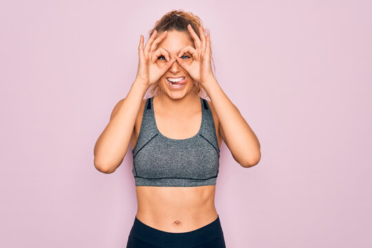 Young beautiful blonde sportswoman with blue eyes doing exercise wearing sportswear doing ok gesture like binoculars sticking tongue out, eyes looking through fingers. Crazy expression.