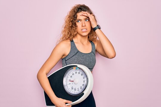 Young beautiful slim sporty woman with blue eyes controlling weight holding weighting machine stressed with hand on head, shocked with shame and surprise face, angry and frustrated. Fear and upset