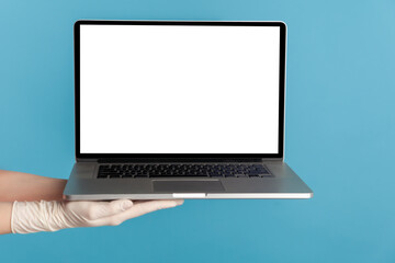 Fototapeta premium Profile side view closeup of human hand in white surgical gloves holding computer laptop and showing empty copy space on monitor screen. indoor, studio shot, isolated on blue background.