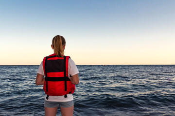 Girl in bright red life jacket stands on seashore with waves, rear view. Rescue equipment for water...