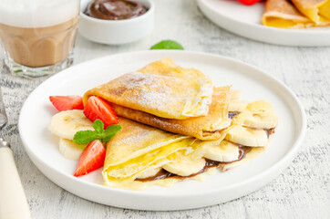 Thin pancakes or crepes with chocolate cream, banana and strawberries on a white plate with a glass of cappuccino on a light wooden background. Horizontal orientation.