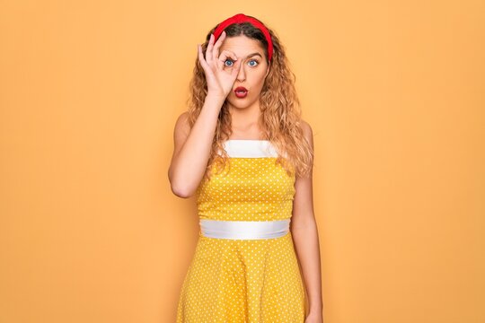 Beautiful blonde pin-up woman with blue eyes wearing diadem standing over yellow background doing ok gesture shocked with surprised face, eye looking through fingers. Unbelieving expression.