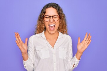 Young beautiful woman with blue eyes wearing casual shirt and glasses over purple background celebrating mad and crazy for success with arms raised and closed eyes screaming excited. Winner concept