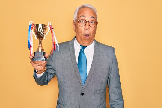 Middle age senior grey-haired business man wearing glasses holding winner trophy scared in shock with a surprise face, afraid and excited with fear expression