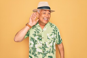 Middle age senior grey-haired man wearing summer hat and floral shirt on beach vacation smiling with hand over ear listening an hearing to rumor or gossip. Deafness concept.